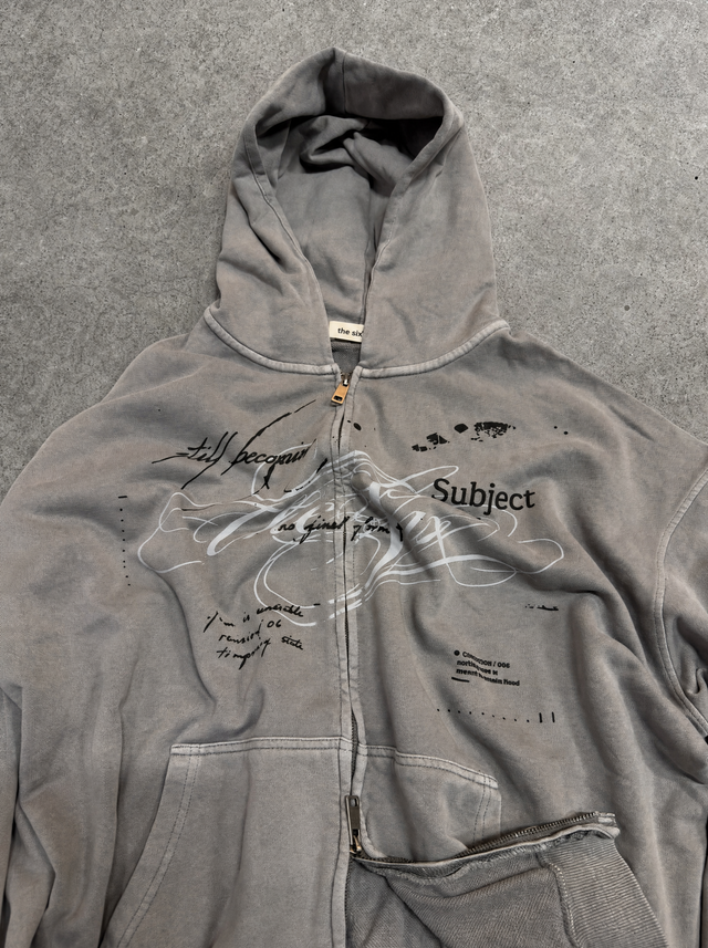 A gray zip-up hoodie with a distressed, washed texture, featuring graphic prints and the word 'Subject' on the front, laid flat on a gray textured background.